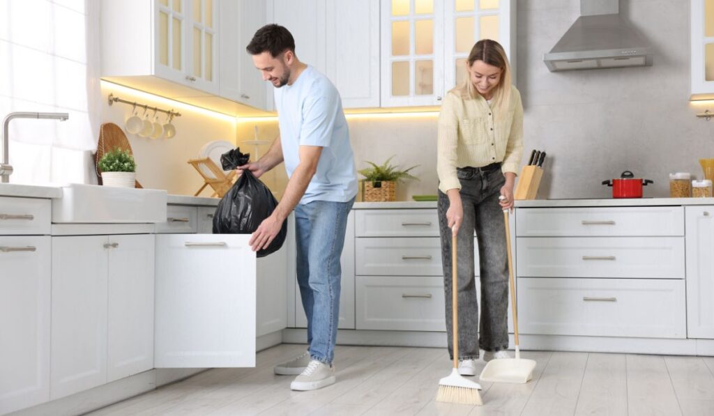 Pareja limpiando cocina, él tira basura, ella barre