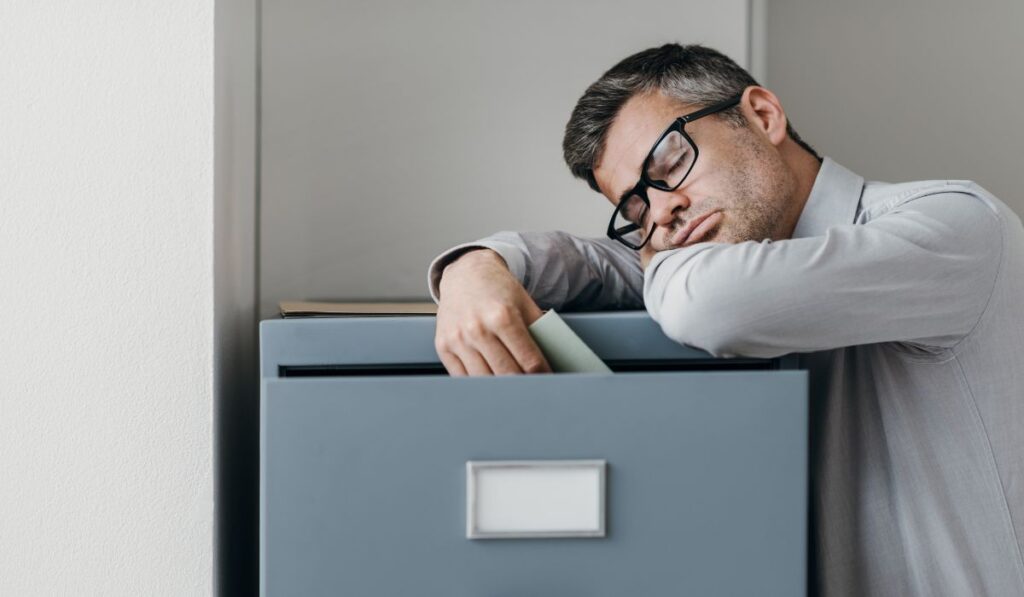 Hombre durmiendo apoyado en un archivador de oficina.