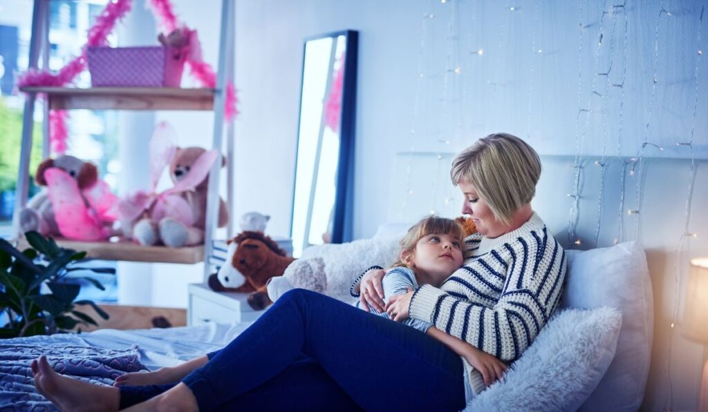 Madre abraza a su hija en la cama, rodeadas de peluches.