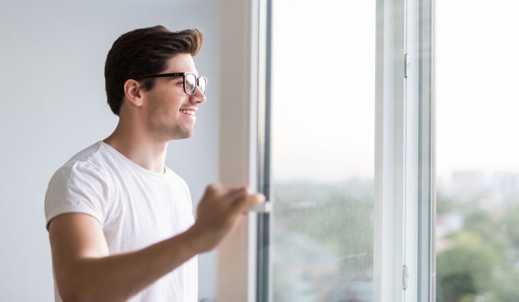 Hombre sonriente con gafas abre una ventana, mirando hacia afuera.