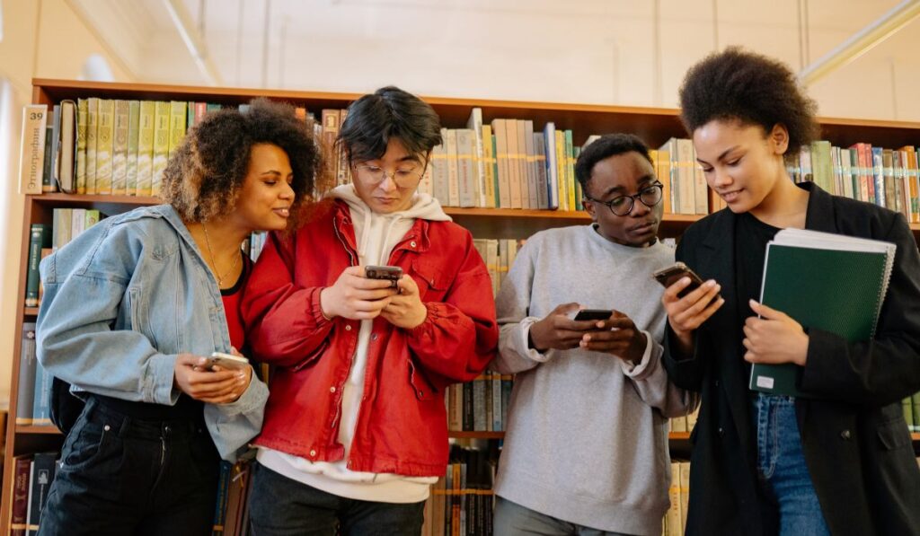 Grupo de estudiantes mirando sus teléfonos frente a estanterías de libros.