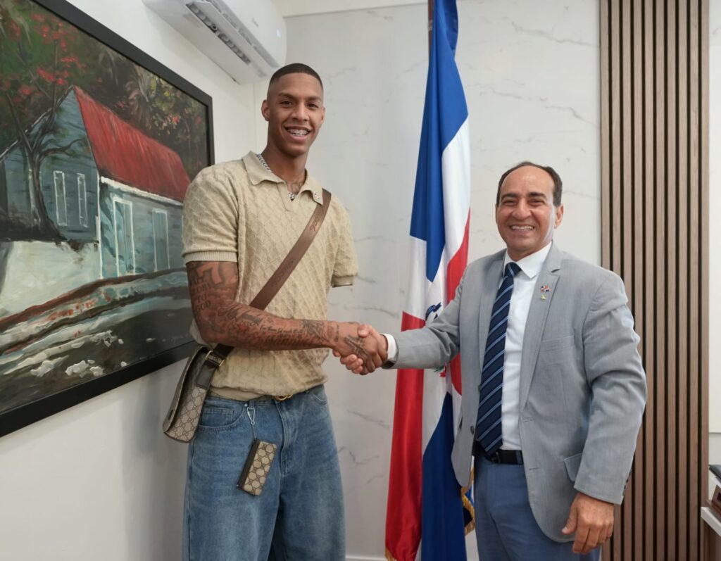 Dos hombres dan la mano frente a la bandera de la República Dominicana.