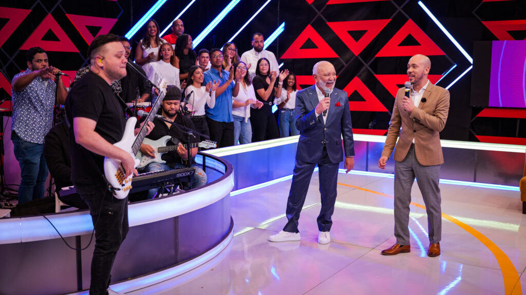 Músicos y presentadores en un escenario de televisión con público.