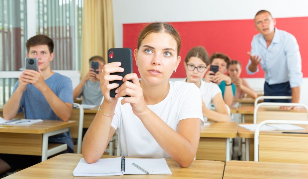Estudiantes distraídos con sus teléfonos en clase mientras el profesor habla.