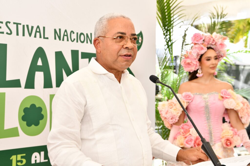 Hombre habla en un atril, detrás una mujer con vestido de flores.