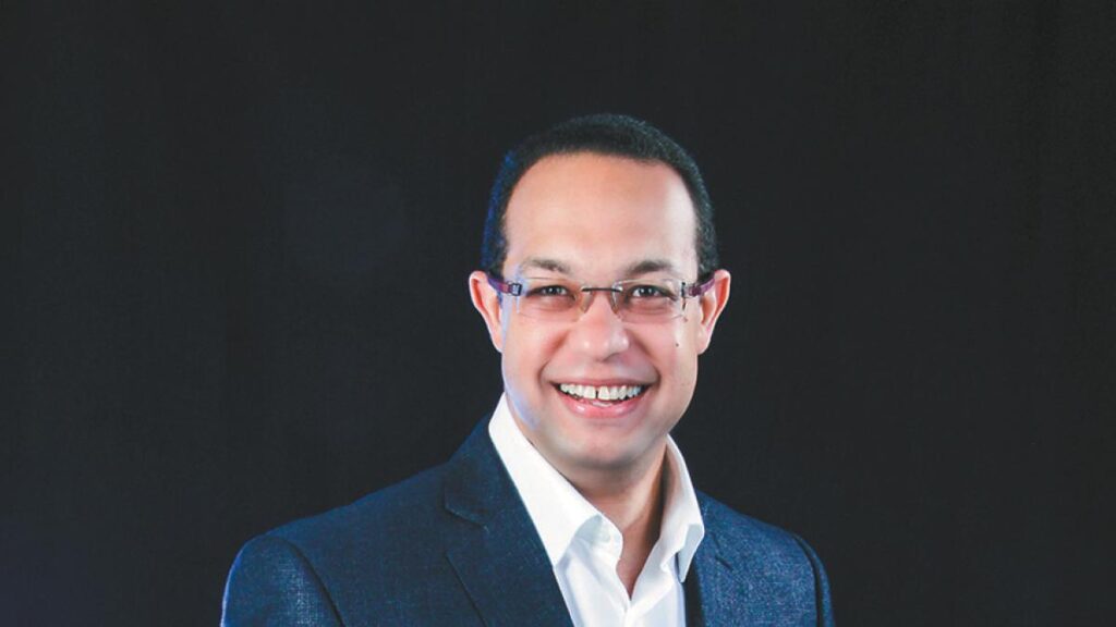Hombre sonriente con gafas y traje oscuro sobre fondo negro.