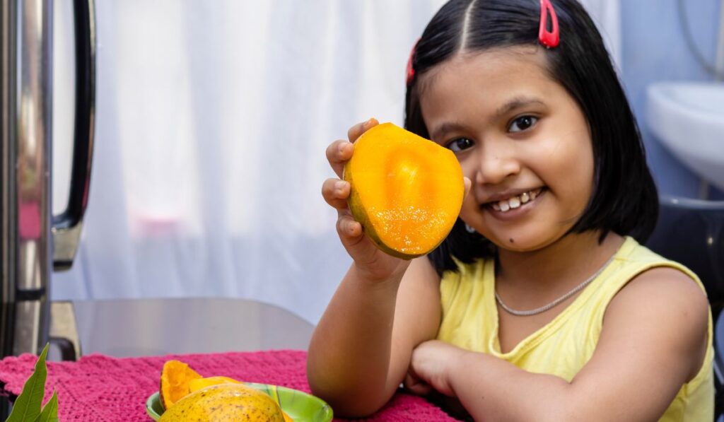 Niña sonriente mostrando la mitad de un mango jugoso.