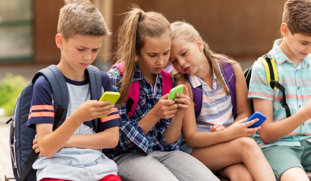 Niños absortos en sus teléfonos móviles, con mochilas, sentados juntos.